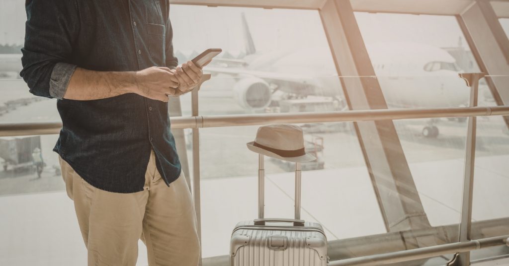 Image of a man holding a phone at an airport, accompanied by a suitcase